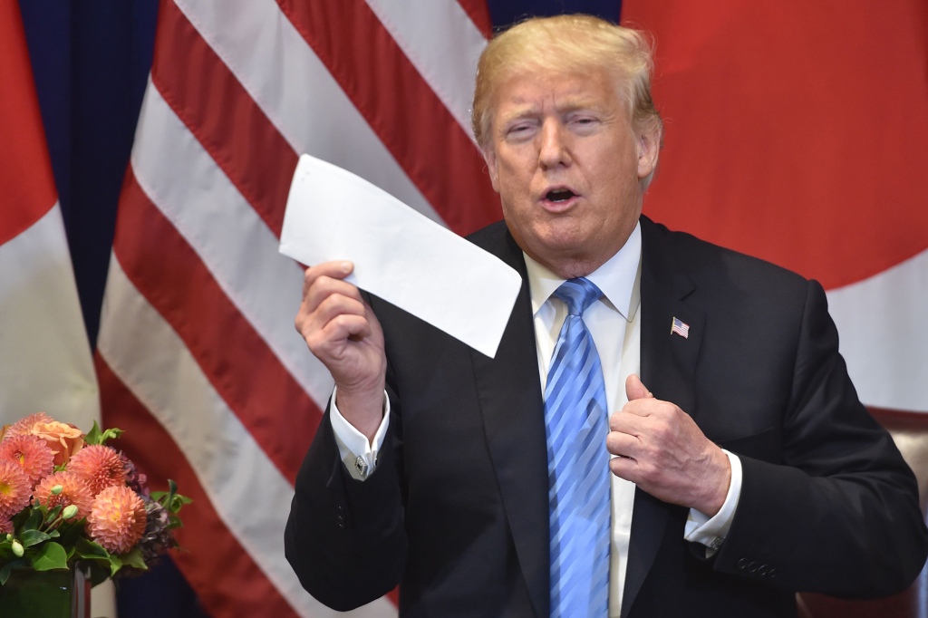 Donald Trump holds up a folded piece of paper with an American flag behind him.