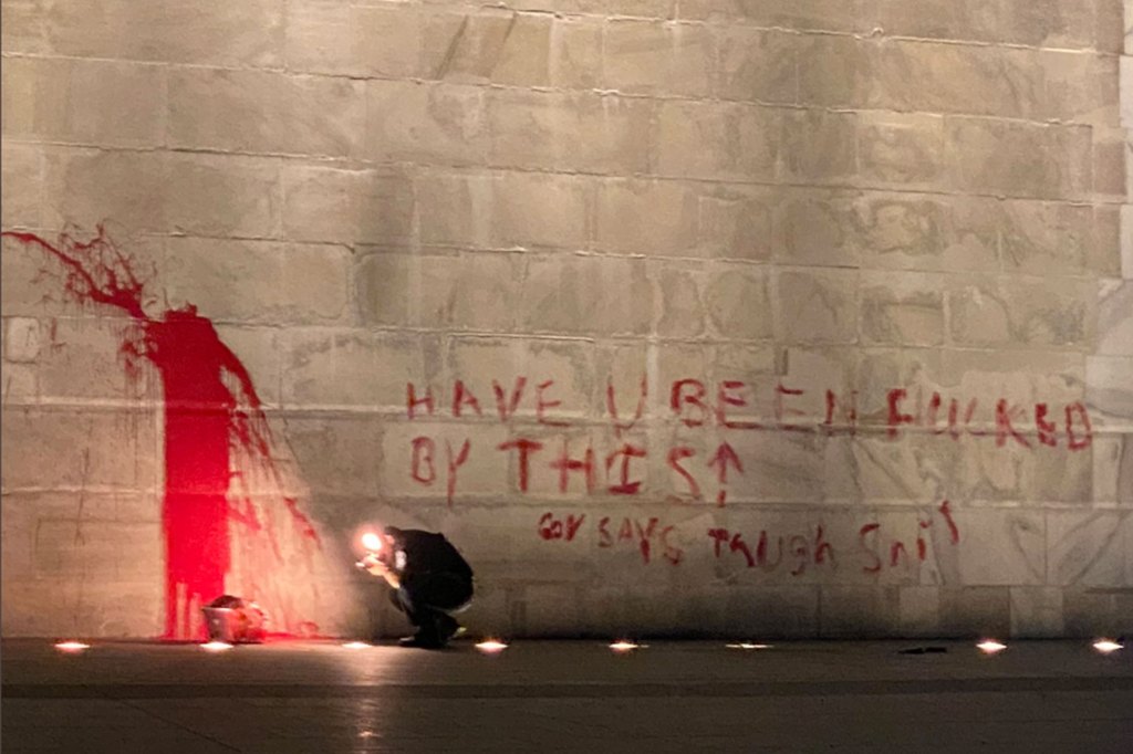 A person crouches to take a photo of red paint splashed on the Washington Monument. The words 